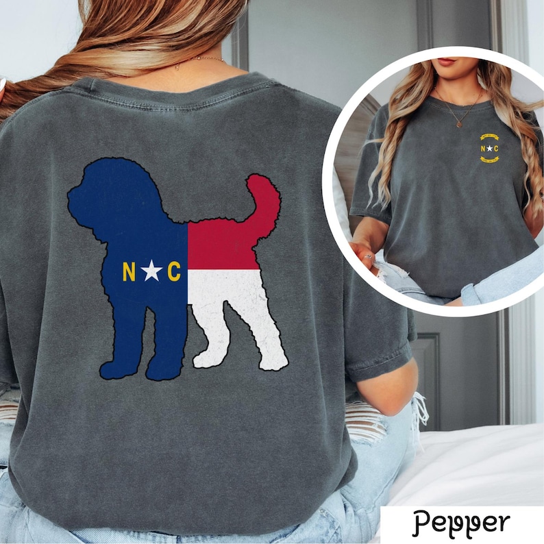 May include: A charcoal grey t-shirt with a distressed look featuring a silhouette of a dog with the North Carolina state flag design. The back of the shirt has the text "N*C" in white. The front of the shirt has a small circle with the text "N*C" in yellow.