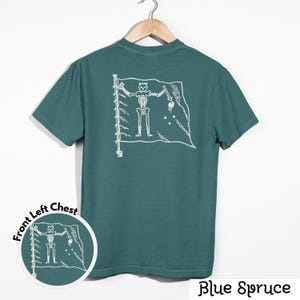 May include: Blue spruce t-shirt with a white skeleton pirate flag design on the back. The shirt has a crew neck and short sleeves. A smaller version of the flag design is on the front left chest. The words "Blue Spruce" are at the bottom.