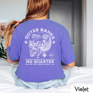 May include: A purple t-shirt with a white graphic of a skull with a compass and anchor inside. The text "Outer Banks NC No Quarter On The Carolina Coast" is printed on the shirt.