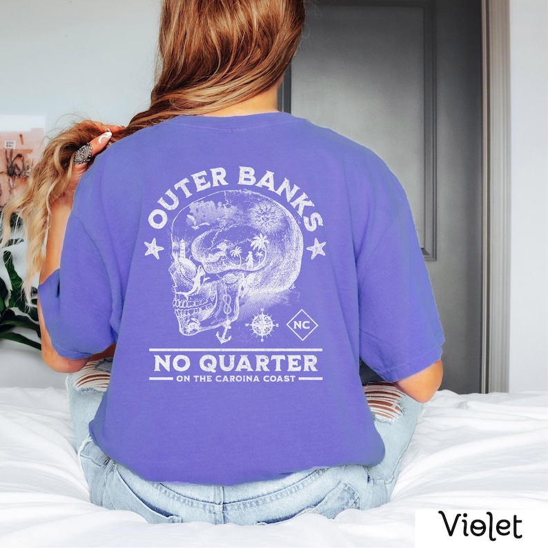 May include: A purple t-shirt with a white graphic of a skull with a compass and anchor inside. The text "Outer Banks NC No Quarter On The Carolina Coast" is printed on the shirt.