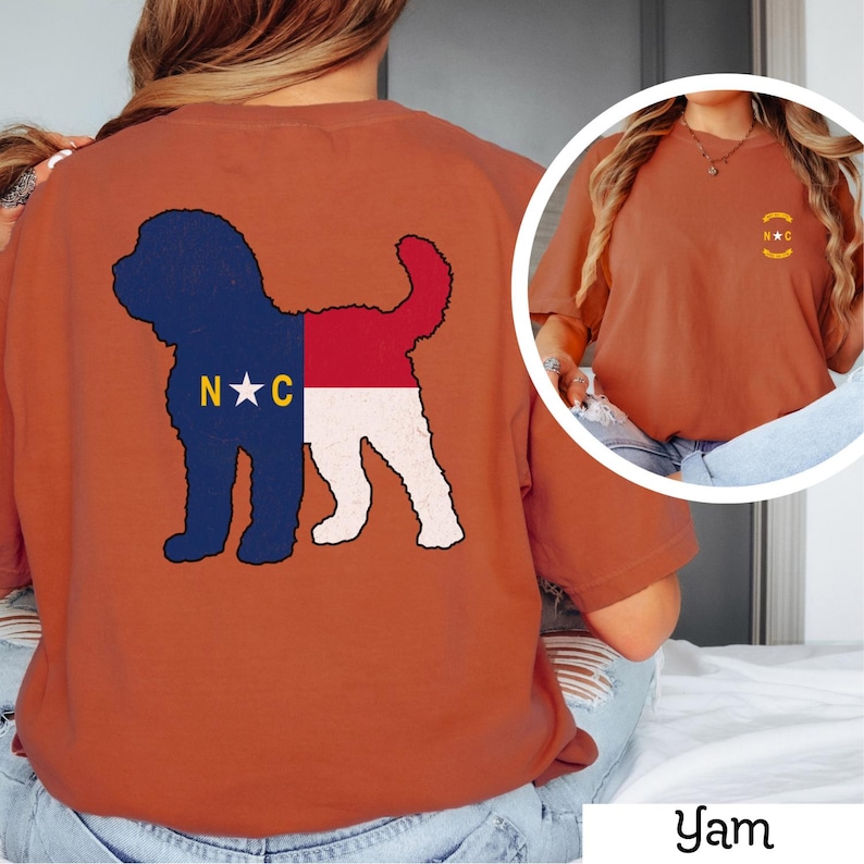 May include: A burnt orange t-shirt with a silhouette of a dog with the North Carolina state flag design on the back. The front of the shirt has a small embroidered design of the North Carolina state flag with the letters "N.C." in gold.