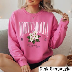May include: Pink sweatshirt with the text "NORTH CAROLINA" in white arched lettering. Below the text is a floral graphic of white dogwood blossoms and the words "the Dogwood State". The sweatshirt has the text "ES TD 17 89".