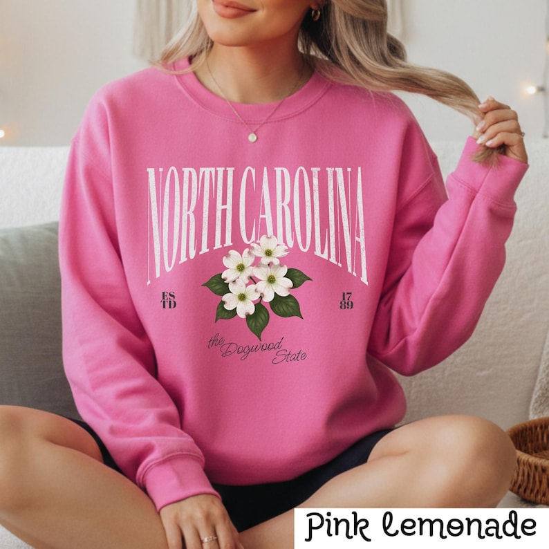May include: Pink sweatshirt with the text "NORTH CAROLINA" in white arched lettering. Below the text is a floral graphic of white dogwood blossoms and the words "the Dogwood State". The sweatshirt has the text "ES TD 17 89".