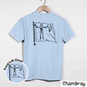 May include: Light blue t-shirt with a black graphic of a skeleton holding a sword and a flag. The flag has a skull and crossbones design. The shirt is hanging on a wooden hanger.