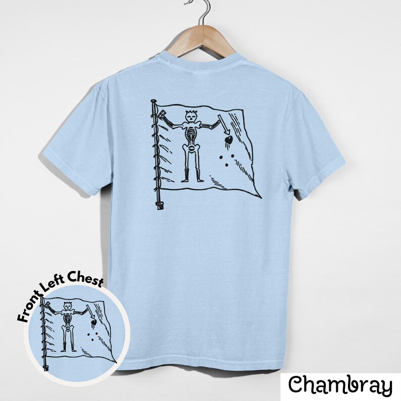 May include: Light blue t-shirt with a black graphic of a skeleton holding a sword and a flag. The flag has a skull and crossbones design. The shirt is hanging on a wooden hanger.