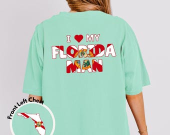 May include: A light mint green t-shirt featuring the text I LOVE MY FLORIDA MAN on the back, with the letters filled by the Florida state flag. A small circular graphic on the lower left shows the outline of Florida with the state seal inside.