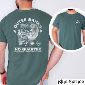 May include: A teal blue t-shirt with a white graphic of a skull with a compass and palm trees inside. The text "Outer Banks No Quarter On The Carolina Coast" is printed below the skull. The t-shirt has a small white square with the letters "NC" on the left chest.