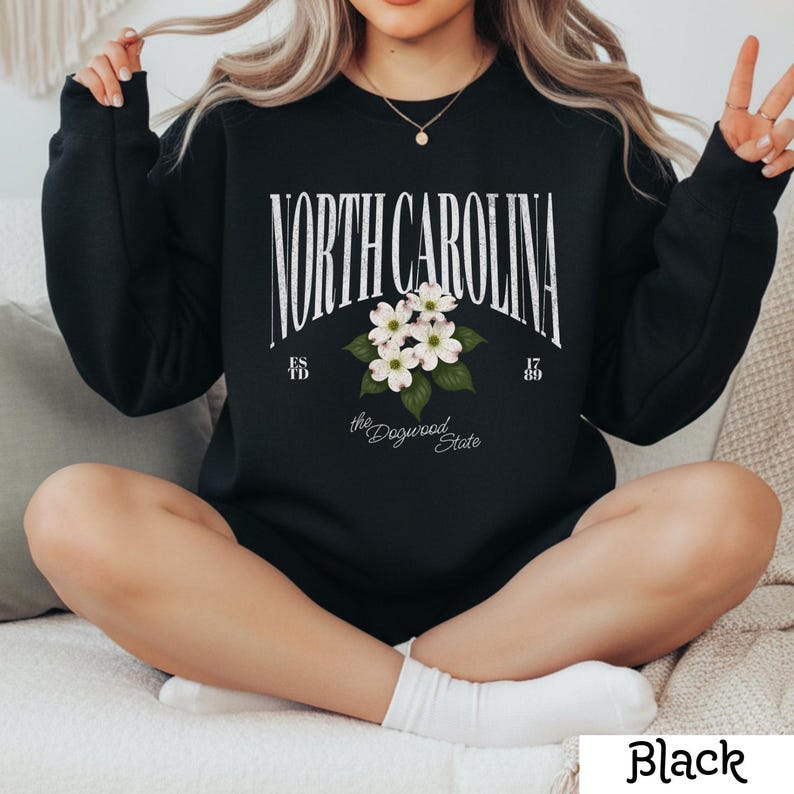 May include: Black sweatshirt featuring the text "NORTH CAROLINA" in a stylized font, with a floral graphic of white dogwood blossoms. Below the text are the words "the Dogwood State" and the numbers "1789" and "ESTD 49".