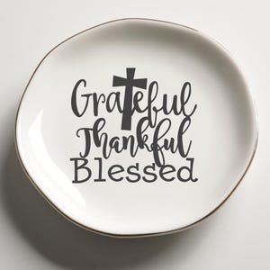 Può includere: Un piatto in ceramica bianca con bordo dorato e forma irregolare. Il piatto presenta le parole "Grateful, Thankful, Blessed" in nero, con una croce sopra la parola "Grateful".
