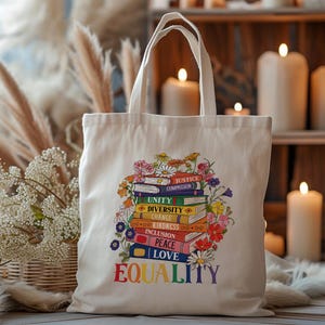 May include: Cream-colored tote bag with a colorful floral and book design. The design includes the words "Equality", "Love", "Peace", "Inclusion", "Kindness", "Change", "Diversity", "Unity", and "Justice". The bag has two handles.