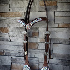 Puede incluir: Una brida de caballo de cuero marrón con intrincados herrajes plateados y brillantes detalles de cristal iridiscente. La testera y las mejilleras están adornadas con cristales de colores. La brida está diseñada para uso ecuestre.
