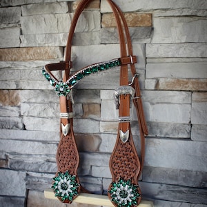 Puede incluir: Una brida de caballo de cuero marrón con intrincados detalles y herrajes plateados. La testera y las mejilleras están adornadas con detalles de cristal verde esmeralda y transparente. La brida está diseñada para uso ecuestre.