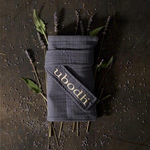 May include: A dark grey wrap with the embroidered word "ubodhi" in gold thread. The wrap is surrounded by sprigs of lavender and loose lavender buds, all arranged on a dark wooden surface.