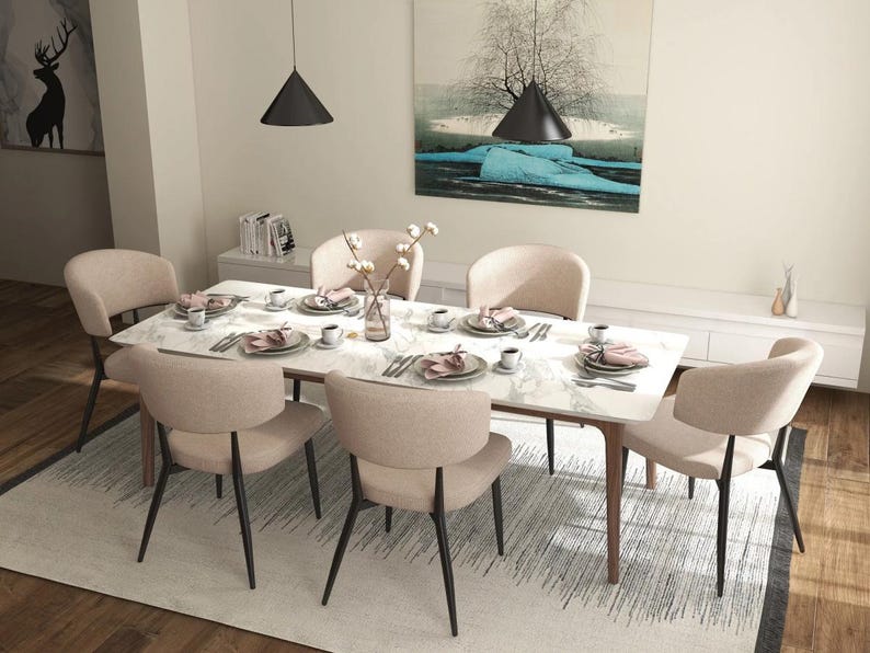 May include: A white marble dining table with six beige chairs. The table is set with place settings, including plates, napkins, and silverware. There is a vase of white flowers in the center of the table.
