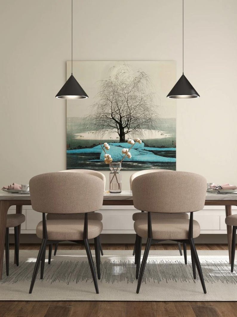 May include: A dining room with a wooden table and six chairs. The chairs are upholstered in a light brown fabric and have black metal legs. Two black pendant lights hang above the table. A large painting of a tree with blue water and white cotton balls hangs on the wall behind the table.