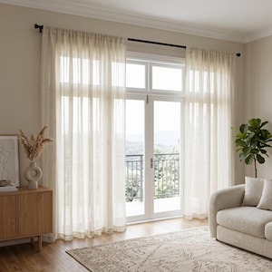 May include: Floor-to-ceiling sheer curtains in a light cream colour, featuring a vertical striped texture. The curtains are hung on a dark metal rod and cover a large glass door, allowing soft light to filter through the semi-transparent fabric.
