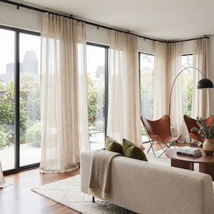 May include: Floor-to-ceiling sheer beige curtains hang from a black metal rod covering large glass sliding doors. The curtains feature a soft, translucent fabric that allows natural light to filter through, creating a bright and airy atmosphere.