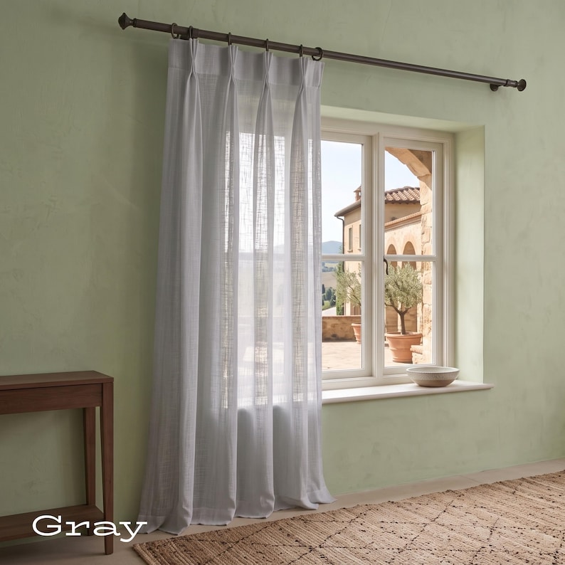 Puede incluir: Una cortina de color gris claro semitransparente con pliegues verticales que cuelga de una barra de metal marr&oacute;n oscuro con anillas. La tela tiene una textura tejida visible que deja pasar la luz, cubriendo una ventana que da al exterior.