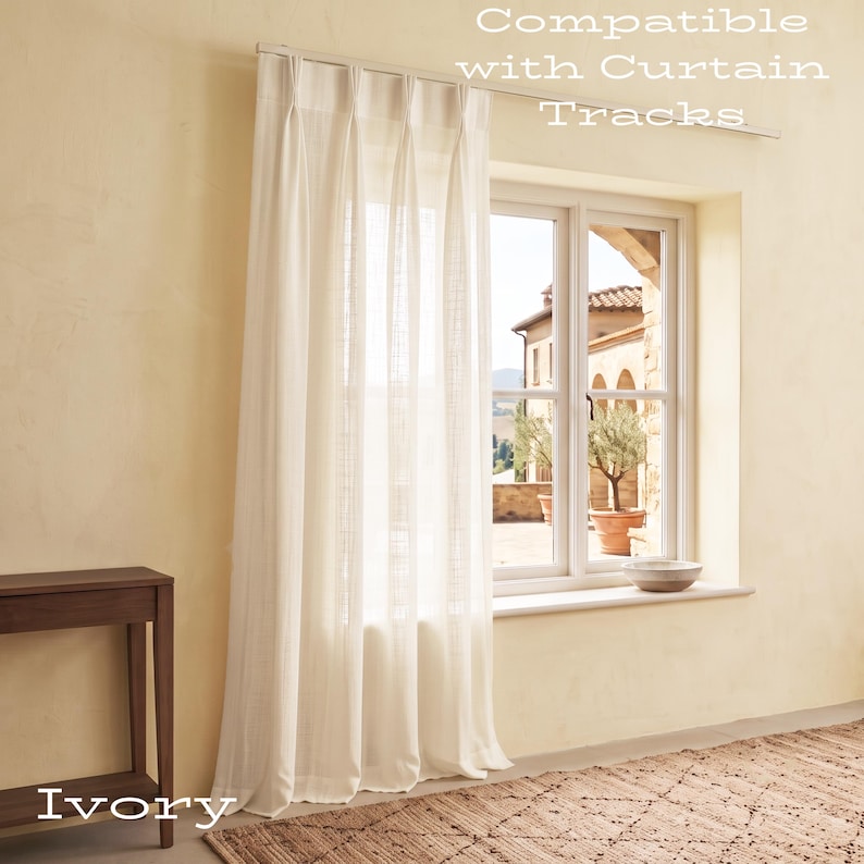 Puede incluir: Una cortina de color marfil con pliegues cuelga de una barra met&aacute;lica sobre una ventana. El texto "Compatible with Curtain Tracks" es visible en la parte superior y la palabra "Ivory" aparece en la parte inferior izquierda.
