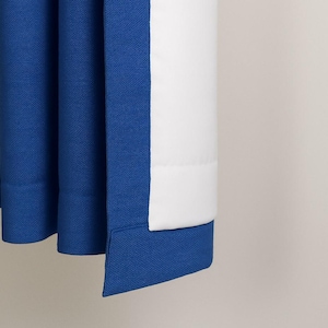 May include: Close-up of a blue and white curtain. The curtain is made of a textured fabric. The blue section has a folded hem. The white section is smooth and appears to be a lining.