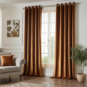 May include: A pair of golden-brown velvet curtains hang from a silver rod, framing a window with sheer white curtains. A matching throw pillow rests on a light-colored sofa. A framed botanical print and a potted plant complete the room's decor.