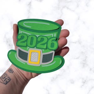 May include: A green St. Patrick's Day hat with the year 2026 in large green numbers. The hat has a grey band with a yellow buckle. The hat is made of paper and is held in a hand.