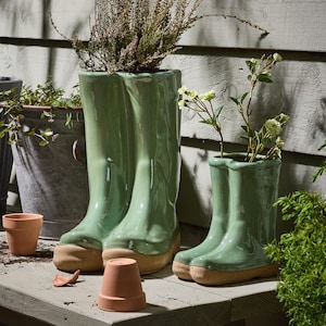 May include: Two green ceramic wellington boot-shaped planters with tan soles, filled with plants. A smaller boot planter and a terracotta pot are also present. The planters are on a wooden surface.