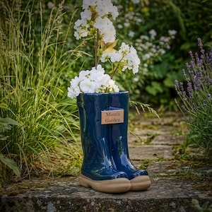 May include: A navy blue ceramic boot vase filled with white flowers. The vase has a tan sole and a wooden plaque that reads "Mum's Garden". The vase is on a stone surface, with greenery in the background. A charming garden ornament.