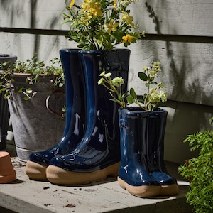 May include: Three navy blue ceramic wellington boot planters with tan soles, filled with yellow and white flowers. The planters are on a wooden surface, with a galvanized metal bucket and greenery in the background.