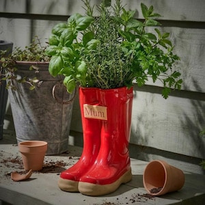 May include: A red ceramic wellington boot planter filled with herbs, with a wooden tag reading "Mum". The boots have a beige sole. Other planters and a broken terracotta pot are in the background, creating a garden scene.