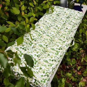 May include: A rectangular outdoor bench cushion with a white background and a green leaf pattern. The cushion is tufted and has a handle on one side. The bench is surrounded by green foliage.