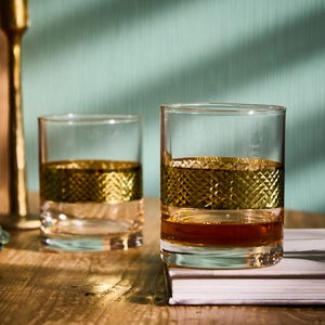 May include: Two clear, lowball glasses with a gold diamond-patterned band around the middle. The glasses are filled with a brown liquid, likely a spirit. The background is a light teal colour.