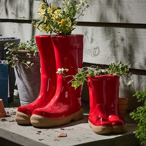 May include: Three glossy red ceramic wellington boot planters with beige soles, filled with plants and flowers. They are set on a wooden surface, with a weathered wood backdrop. A small terracotta pot is visible.