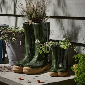 May include: Green ceramic wellington boot planters with tan soles. The planters are filled with plants. A small, medium, and large boot planter are displayed on a wooden surface. Other planters are in the background.