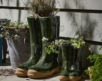 May include: Green ceramic wellington boot planters with tan-colored soles. The planters are filled with plants and greenery. A small, medium, and large boot planter are displayed on a wooden surface. Other planters are in the background.