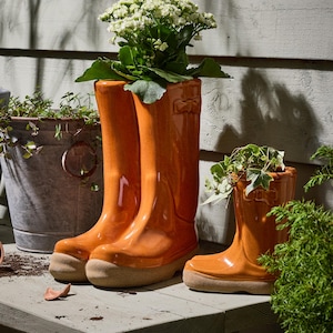 May include: Two orange ceramic wellington boot planters, one large and one small, filled with plants. The large planter is a pair of boots, and the small planter is a single boot. The planters are on a wooden surface.