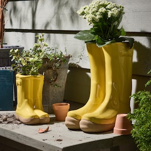 May include: Two bright yellow ceramic wellington boot planters, one large and one small, filled with greenery and flowers. The boots have tan-coloured soles and are set on a weathered wooden surface, with a small terracotta pot nearby.