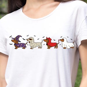 Puede incluir: Camiseta blanca con cuatro perros salchicha disfrazados para Halloween: una bruja, una momia, un diablo y un fantasma. El perro bruja lleva una túnica y un sombrero morados. Murciélagos negros dispersos.