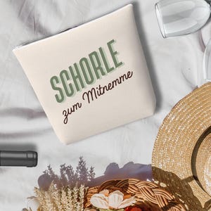 Op de afbeelding: Een picknickscène met een strohoed, wijnglazen en een fles wijn. Een gevlochten mand is gevuld met bloemen. Een canvas tas met de tekst "SCHORLE zum Mitnemme" is ook aanwezig.