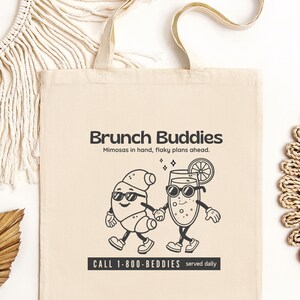 Puede incluir: Bolso tote de lona beige con el texto "Brunch Buddies" y una ilustración de dibujos animados de un mimosa y un personaje de huevo tomados de la mano. También se incluye el texto "Mimosas in hand, flaky plans ahead". La parte inferior de la bolsa tiene el texto "CALL 1-800-BEDDIES served daily."