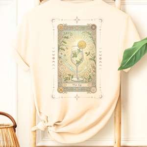 Op de afbeelding: Een beige T-shirt met een afbeelding van een glas limonade met een schijfje citroen en een takje munt. De afbeelding is omgeven door een decoratief kader met de tekst "The Sip".