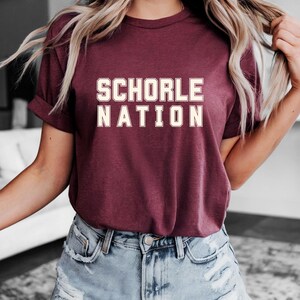 Può includere: T-shirt bordeaux con la scritta "SCHORLE NATION" in grassetto, color bianco sporco. La maglietta ha maniche corte e girocollo. La modella indossa pantaloncini di jeans azzurri. Lo stile generale è casual.