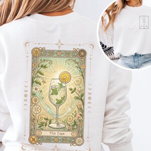 Peut inclure: Sweat-shirt blanc avec un motif de carte de tarot dans le dos. La carte représente un cocktail avec une tranche de citron, de la menthe et des bulles. La carte est encadrée d'éléments floraux et célestes, avec les mots "The Ease" en bas.