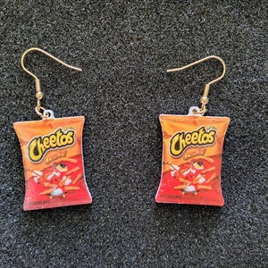 May include: A pair of gold hoop earrings with a red and orange Cheeto bag charm. The bag has the text "Cheetos Crunchy" printed on it.