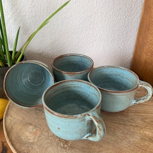 Könnte beinhalten: Ein Set aus vier hellblauen Keramikbechern mit gesprenkelter Oberfläche und braunen Rändern. Die Becher haben eine abgerundete Form mit einem stabilen Henkel an der Seite. Das Innere jedes Bechers zeigt eine glatte Glasur mit feinen Strukturen.