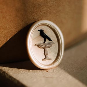 May include: An oval-shaped wax seal with a black raven perched on a pedestal. The seal is off-white with a subtle pearlescent sheen. The background is a warm, neutral tone, with shadows adding depth.