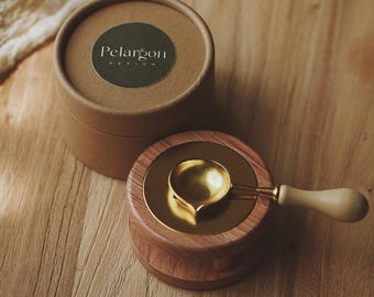 Puede incluir: Un calentador de cera de madera con una cuchara dorada y una caja redonda de cartón con el texto "Pelargon Design" en una etiqueta verde. El calentador está sobre una superficie de madera.