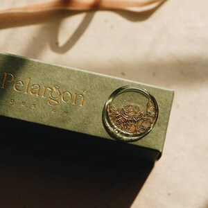 May include: A green box with the text "Pelargon Design" on the top. A clear wax seal with a gold floral design is on the box.