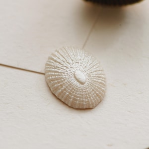 May include: A cream-coloured wax seal with a textured, shell-like design. A gold-coloured metal stamp is positioned above the seal. The seal is on light-coloured paper, suggesting stationery or craft supplies.