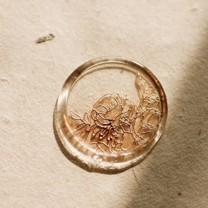 May include: A clear wax seal with a floral design in gold. The seal is round and has a raised edge.
